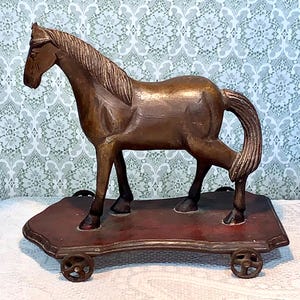 May include: A brown wooden horse sculpture with a detailed mane and tail, mounted on a rectangular, reddish-brown base with small wheels. The horse is posed with one leg raised, giving it a dynamic appearance.