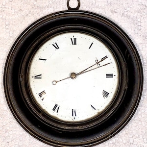 May include: Circular antique wall clock with a black frame and a white face. The clock has black Roman numerals and slender hands. A small metal loop is at the top for hanging. The clock's diameter is approximately 20 cm.