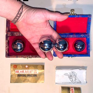 Op de afbeelding: Een hand houdt twee glanzende, zilveren metalen ballen vast, met een derde in een blauw-rode doos. De doos is open en onthult een rode binnenkant. Twee verpakkingen met Chinese tekst en de woorden "IRON BALL DIRECTION" zijn ook zichtbaar.