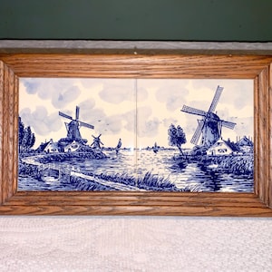 Peut inclure: Une œuvre d'art encadrée représentant un paysage de moulins à vent hollandais en bleu et blanc. L'œuvre est composée de deux carreaux avec un paysage détaillé de moulins à vent, d'eau et de maisons. Le cadre est en bois.