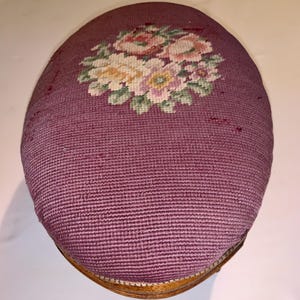 Pode incluir: Banqueta oval com tampo de tecido malva. O tampo apresenta um desenho floral em ponto de agulha em tons de rosa, amarelo e verde. A base é de madeira. Uma peça vintage.