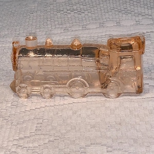 May include: A vintage, peach-coloured glass train figurine. The train features detailed moulding of the engine, carriages, and wheels. The glass is transparent, allowing light to pass through. This decorative piece is a collectible item.