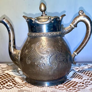 May include: A silver teapot with an ornate design and a floral pattern. The teapot has a lid with a finial and a handle.
