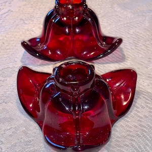 May include: Two ruby red glass candle holders. Each candle holder has a flower-like design with curved petals and a central cup for a candle. The glass has a glossy finish, reflecting light.