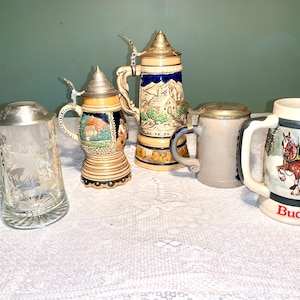 May include: A selection of beer steins, showcasing diverse styles and materials. Includes a clear glass stein with a metal lid, ceramic steins with detailed artwork, and a Budweiser stein with a horse-drawn carriage scene.