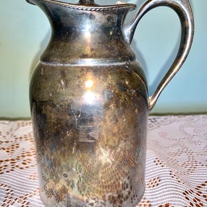 Könnte beinhalten: Eine silberne Kanne mit Deckel und Henkel. Die Kanne hat eine strukturierte Oberfläche und ist leicht angelaufen.