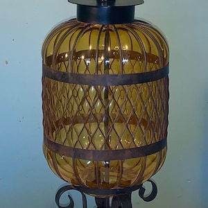 May include: A vintage amber glass lamp with a decorative metal cage and base. The lamp features a cylindrical glass shade with a metal cage design and a black metal base. The lamp is approximately 61 cm tall.