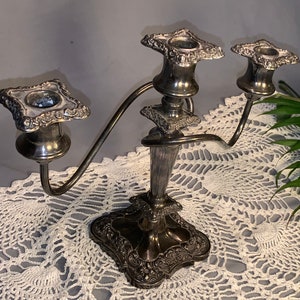 May include: A silver three-armed candelabra with ornate details and a scrolled design. The candelabra is sitting on a white crocheted doily.
