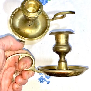 Peut inclure: Deux bougeoirs en laiton antique au design simple. L'un a une poignée et une base ronde. L'autre a une base ronde et une tige haute et fine.