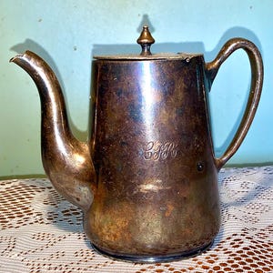 Op de afbeelding: Een verzilverde theepot met een aangetast oppervlak en een klein knopje op de deksel. De theepot heeft een handvat en een tuit. De letters "C.F.R." zijn gegraveerd op de zijkant van de theepot.