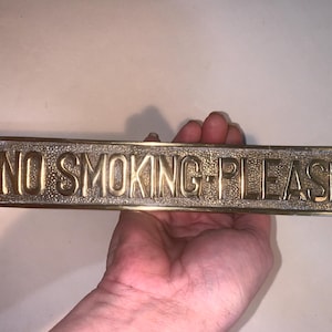 May include: A rectangular brass sign with the words "NO SMOKING PLEASE" embossed on its surface. The sign is held in a hand, and the text is in capital letters. The sign has a textured background.