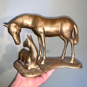 Peut inclure: Une sculpture en laiton d'une jument et de son poulain. La jument debout baisse la tête vers le poulain, qui est couché. La sculpture est sur une base qui ressemble à de l'herbe.