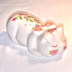 Może przedstawiać: Biała ceramiczna figurka świni w pozycji śpiącej, ozdobiona różowymi kwiatowymi akcentami i pasującą kokardą. Świnia ma zamknięte oczy i mały różowy nos, z kwiatowymi detalami na ciele. Przedmiot dekoracyjny.