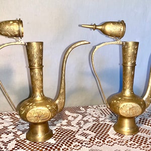 Puede incluir: Dos jarras de metal dorado adornadas con diseños intrincados y tapas. Las jarras tienen un cuerpo redondo y un pico largo y curvo.