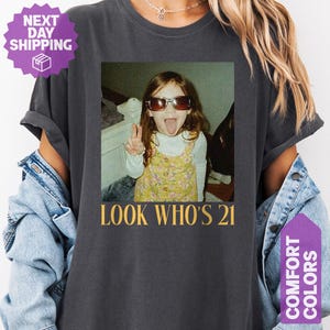 May include: Dark gray t-shirt with a photo print featuring a young person wearing sunglasses and sticking out their tongue. The text "LOOK WHO'S 21" is printed below the photo in gold.