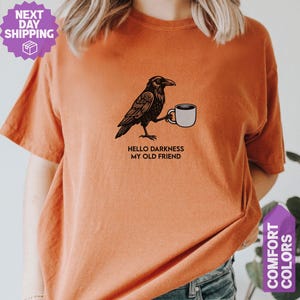 May include: A burnt orange short-sleeve t-shirt featuring a black graphic illustration of a raven holding a white coffee mug. Below the raven, the text reads, "HELLO DARKNESS MY OLD FRIEND" in black capital letters. The shirt has a crew neckline.