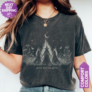 May include: A charcoal gray short-sleeve t-shirt featuring a light-colored graphic of three figures in pointed hats holding hands in a field of wildflowers. Above them is a crescent moon and stars. The text "girls will be girls" is printed below.