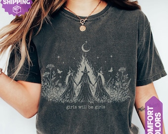 May include: A charcoal gray short-sleeve t-shirt featuring a light-colored graphic of three figures in pointed hats holding hands in a field of wildflowers. Above them is a crescent moon and stars. The text "girls will be girls" is printed below.