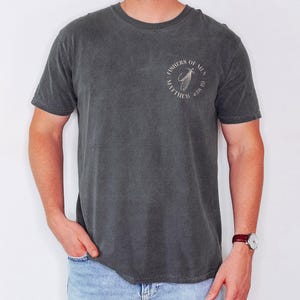 Op de afbeelding: Donkergrijs T-shirt met een ronde hals en korte mouwen. Het shirt heeft een cirkelvormige afbeelding op de linkerbovenborst met de tekst "FISHERS OF MEN" en "MATTHEW 4:18-19". Het shirt is gemaakt van een zacht, comfortabel materiaal.