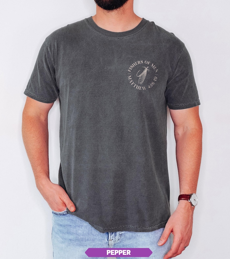 Op de afbeelding: Donkergrijs T-shirt met een ronde hals en korte mouwen. Het shirt heeft een cirkelvormige afbeelding op de linkerbovenborst met de tekst "FISHERS OF MEN" en "MATTHEW 4:18-19". Het shirt is gemaakt van een zacht, comfortabel materiaal.