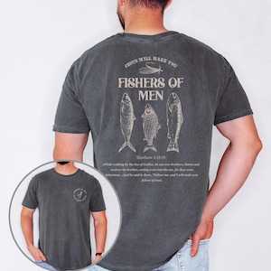 Op de afbeelding: Pepperkleurig t-shirt met een grafisch ontwerp op de achterkant met drie visillustraties en de tekst "JESUS WILL MAKE YOU FISHERS OF MEN" met een bijbelvers. De voorkant van de shirt heeft ook een kleine afbeelding.