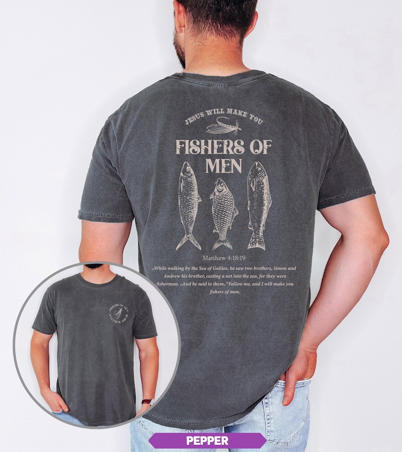 Op de afbeelding: Pepperkleurig t-shirt met een grafisch ontwerp op de achterkant met drie visillustraties en de tekst "JESUS WILL MAKE YOU FISHERS OF MEN" met een bijbelvers. De voorkant van de shirt heeft ook een kleine afbeelding.