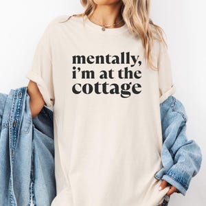 May include: Ivory-colored t-shirt with the text "mentally, I'm at the cottage" in black. The shirt is a relaxed fit. A denim jacket is draped over the shoulders.