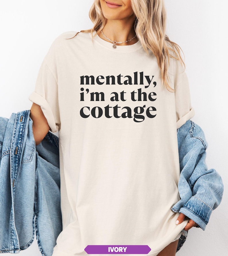 May include: Ivory-colored t-shirt with the text "mentally, I'm at the cottage" in black. The shirt is a relaxed fit. A denim jacket is draped over the shoulders.