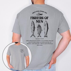 Op de afbeelding: Grijze t-shirt met de tekst "JESUS WILL MAKE YOU FISHERS OF MEN" en drie visillustraties. De achterkant van de shirt bevat ook een bijbelvers uit Matthe&uuml;s 4:18-19. De shirt is een casual, comfortabele stijl.