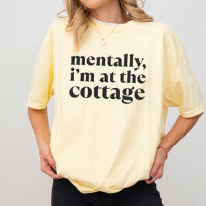 May include: A light yellow t-shirt with the text "mentally, I'm at the cottage" in black. The shirt is a relaxed fit and has short sleeves. The bottom of the shirt is slightly gathered. The word "BANANA" is in a purple rectangle at the bottom.