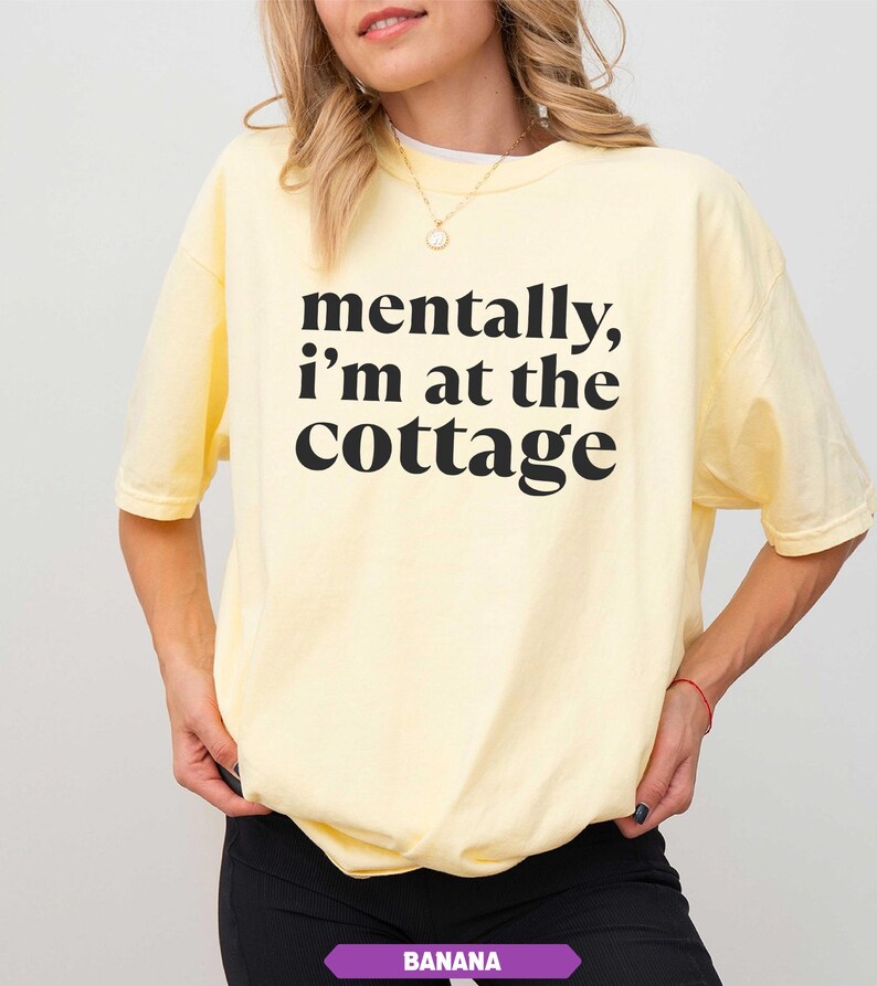 May include: A light yellow t-shirt with the text "mentally, I'm at the cottage" in black. The shirt is a relaxed fit and has short sleeves. The bottom of the shirt is slightly gathered. The word "BANANA" is in a purple rectangle at the bottom.