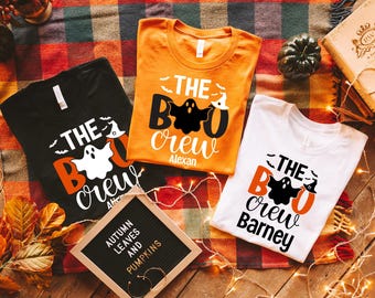 May include: Three Halloween-themed t-shirts in black, orange, and white, each with a unique "The Boo Crew" design featuring a ghost and bat graphics. A sign reads "Autumn Leaves and Pumpkins."