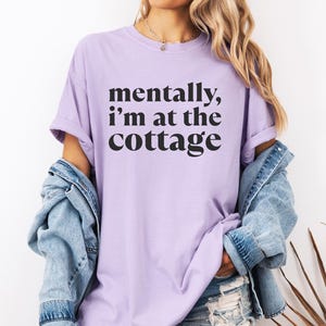 May include: A light purple t-shirt with the text "mentally, I'm at the cottage" in black. The shirt has short sleeves and a relaxed fit. The word "ORCHID" is printed on a purple banner at the bottom of the image.