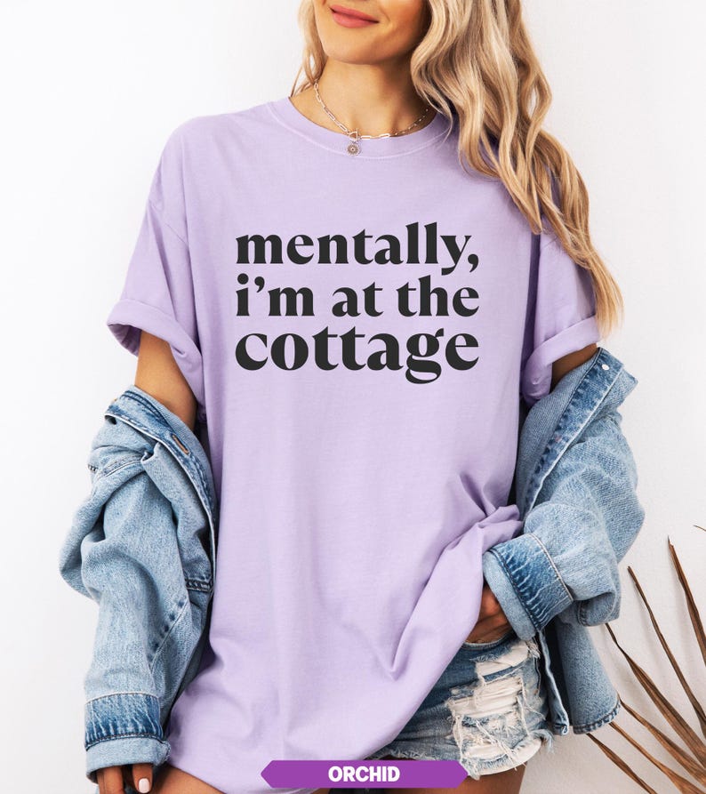 May include: A light purple t-shirt with the text "mentally, I'm at the cottage" in black. The shirt has short sleeves and a relaxed fit. The word "ORCHID" is printed on a purple banner at the bottom of the image.