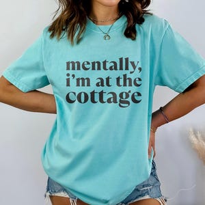 May include: A turquoise t-shirt with the phrase "mentally, I'm at the cottage" printed in dark brown. The shirt has short sleeves and a relaxed fit. The bottom of the shirt has the words "CHALKY MINT" in a purple rectangle.