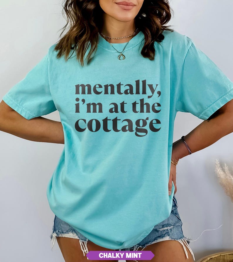 May include: A turquoise t-shirt with the phrase "mentally, I'm at the cottage" printed in dark brown. The shirt has short sleeves and a relaxed fit. The bottom of the shirt has the words "CHALKY MINT" in a purple rectangle.