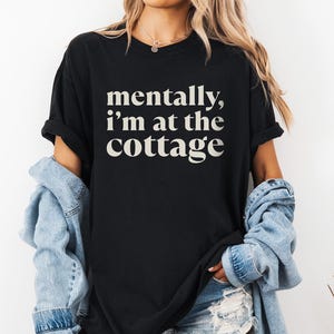 May include: Black t-shirt with the text "mentally, I'm at the cottage" in a white, serif font. The shirt has short sleeves and a relaxed fit. The model is wearing a denim jacket and shorts.