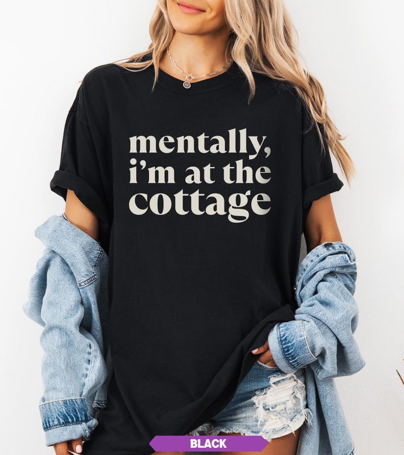 May include: Black t-shirt with the text "mentally, I'm at the cottage" in a white, serif font. The shirt has short sleeves and a relaxed fit. The model is wearing a denim jacket and shorts.