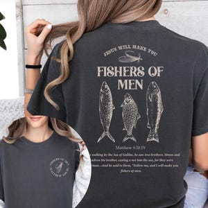 Op de afbeelding: Donkergrijs T-shirt met de tekst "JESUS WILL MAKE YOU FISHERS OF MEN" en drie visillustraties. De tekst "Matthew 4:18:19" staat ook op het shirt. Het shirt heeft de kleur Pepper.