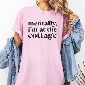 May include: A light pink t-shirt with the text "mentally, I'm at the cottage" in black. The word "BLOSSOM" is printed in a purple rectangle at the bottom. A denim jacket is draped over the shoulders.