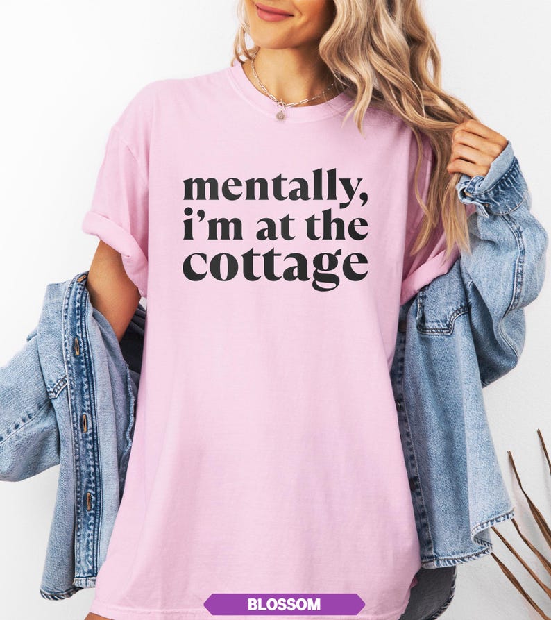May include: A light pink t-shirt with the text "mentally, I'm at the cottage" in black. The word "BLOSSOM" is printed in a purple rectangle at the bottom. A denim jacket is draped over the shoulders.