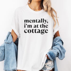 May include: White t-shirt with the phrase "mentally, I'm at the cottage" in black lettering. The model is wearing a light blue denim jacket. The shirt has short sleeves and a relaxed fit. The bottom of the shirt is visible.