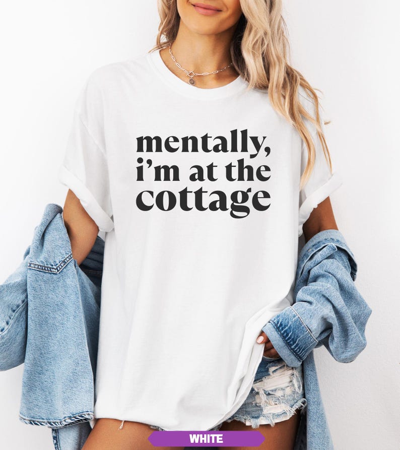 May include: White t-shirt with the phrase "mentally, I'm at the cottage" in black lettering. The model is wearing a light blue denim jacket. The shirt has short sleeves and a relaxed fit. The bottom of the shirt is visible.