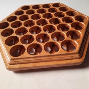 May include: A hexagonal light brown wooden organizer featuring a grid of thirty-seven circular cutouts arranged in a honeycomb pattern. The base has a tiered edge design, and the natural wood grain is visible across the smooth, polished surface.