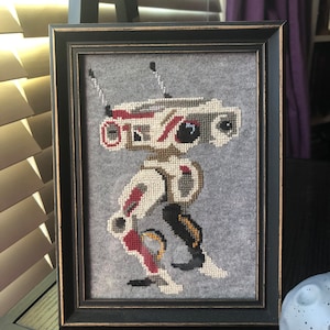 May include: A framed cross-stitch of a white and red robot with black accents. The robot is standing on a grey background. The frame is black with a distressed finish.