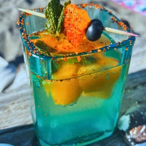 Op de afbeelding: Een turquoise cocktail in een vierkant glas, gegarneerd met munt, een sinaasappelschijfje en een donkere olijf op een tandenstoker. De rand is bedekt met een rood-oranje kruid. De drank bevat sinaasappelschijfjes.