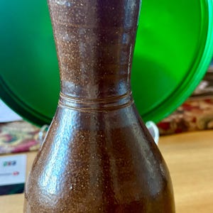 May include: A tall, dark brown ceramic vase with a glossy, speckled finish. The vase features a narrow neck that flares slightly at the top and a rounded, bulbous base. Two distinct horizontal bands are etched into the neck and the lower body.