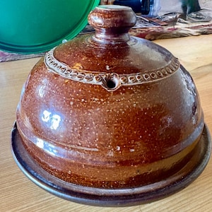 May include: A round, brown glazed ceramic cheese dome with a matching base plate. The dome features a central knob handle and a decorative embossed band around the middle, which includes a small, circular vent hole near the top edge.