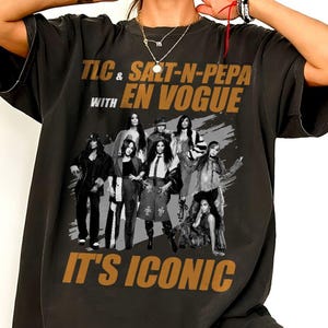 May include: A black short-sleeve t-shirt featuring a graphic print of the musical groups TLC, Salt-N-Pepa, and En Vogue. The text reads TLC & SALT-N-PEPA WITH EN VOGUE and IT'S ICONIC in gold lettering above and below the black and white group photo.