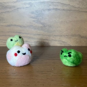 May include: Three felt frog plushies. Two are green and one is pink, white, and green. The green frogs have black eyes and a black mouth. The pink, white, and green frog has black eyes and a black smile.
